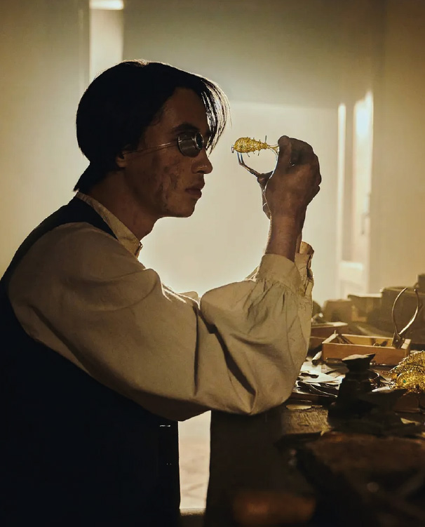 A man with round glasses and antique clothing carefully examines a golden figure in a workshop dimly lit by a soft light.