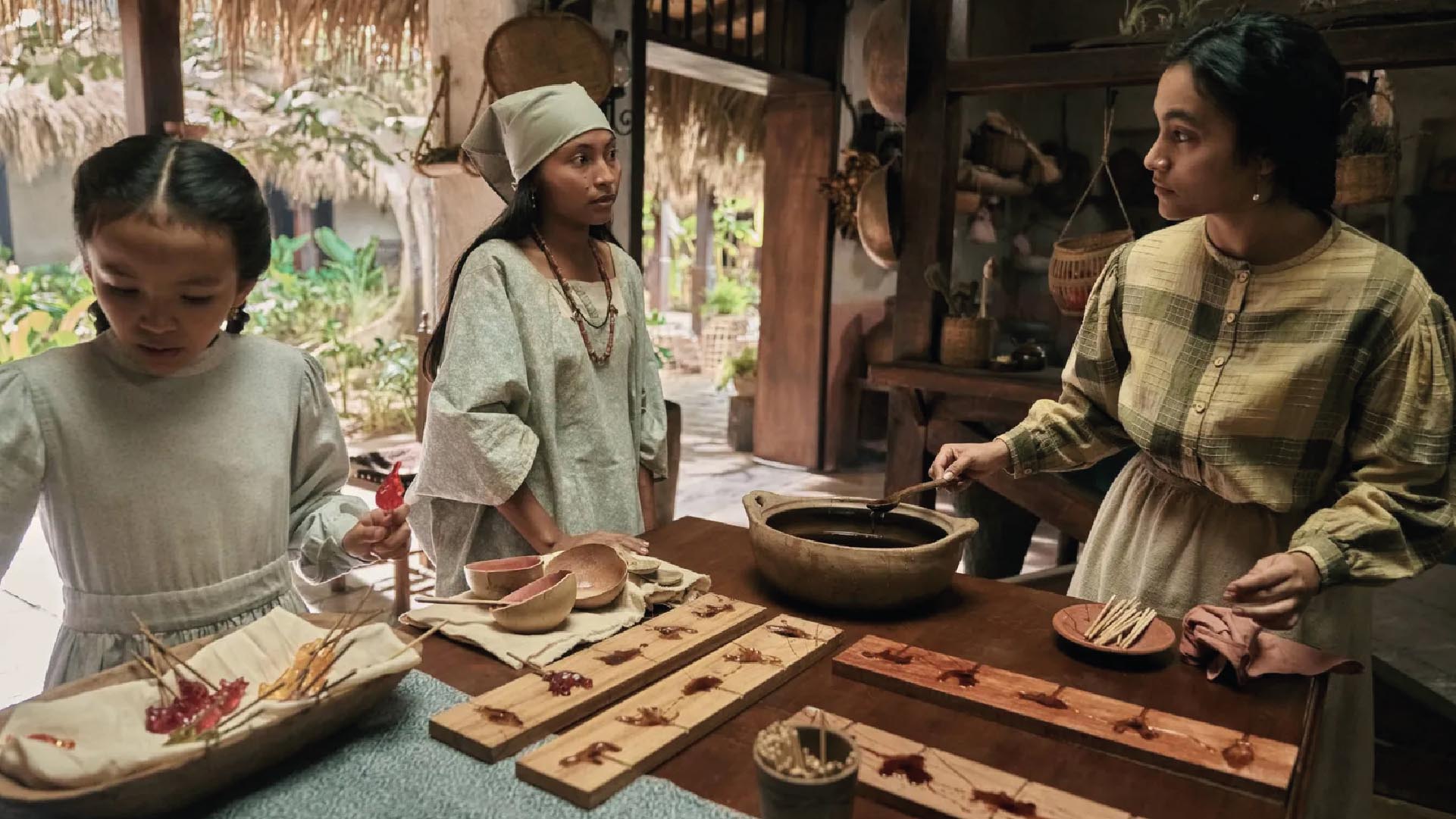 Scene in a rustic kitchen where three women of different ages prepare traditional sweets.