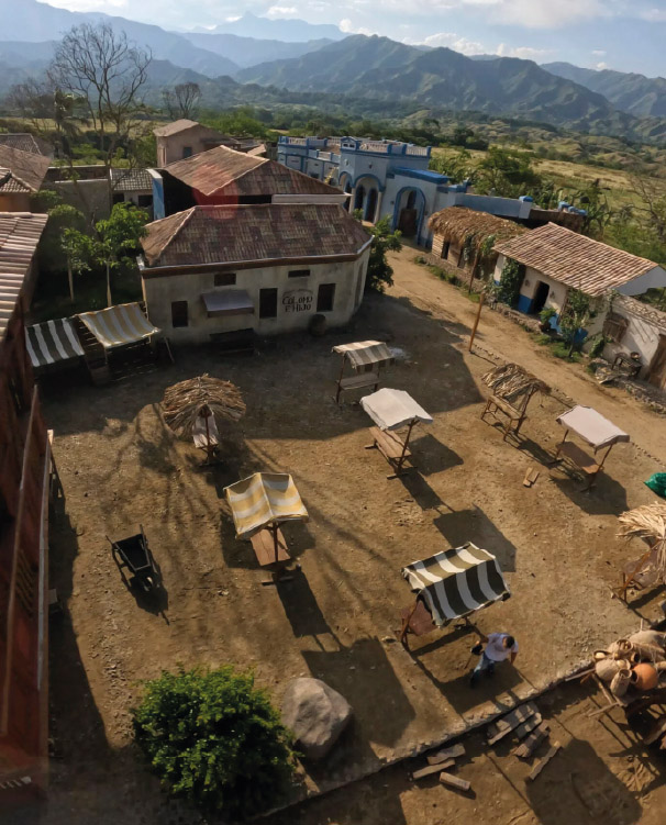 Aerial view of an ancient town with an open-air market, featuring wooden stalls with palm-thatched roofs and striped canvas awnings.