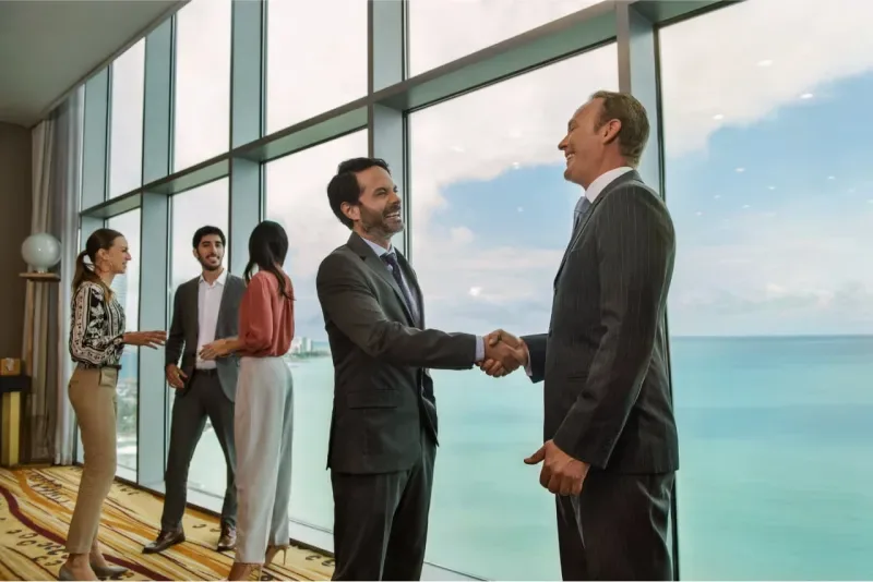 Two businessmen shaking hands in front of a panoramic window overlooking the sea.